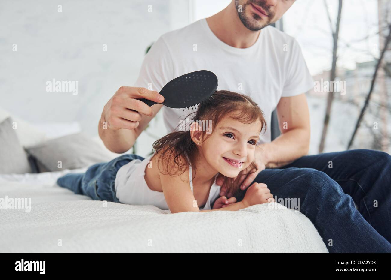 Father makes haircut for his daughter by using comb at home on bed Stock Photo