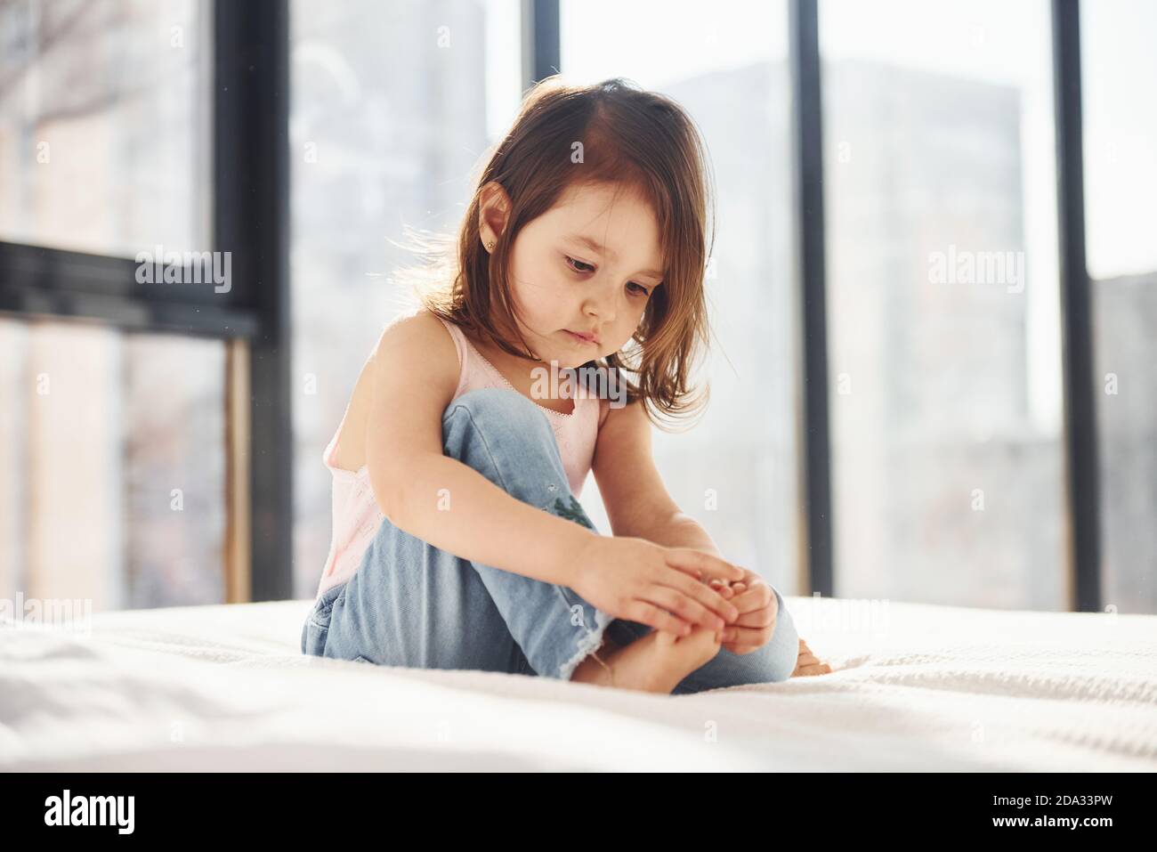Cute little girl in casual clothes sits on bed alone at daytime Stock Photo