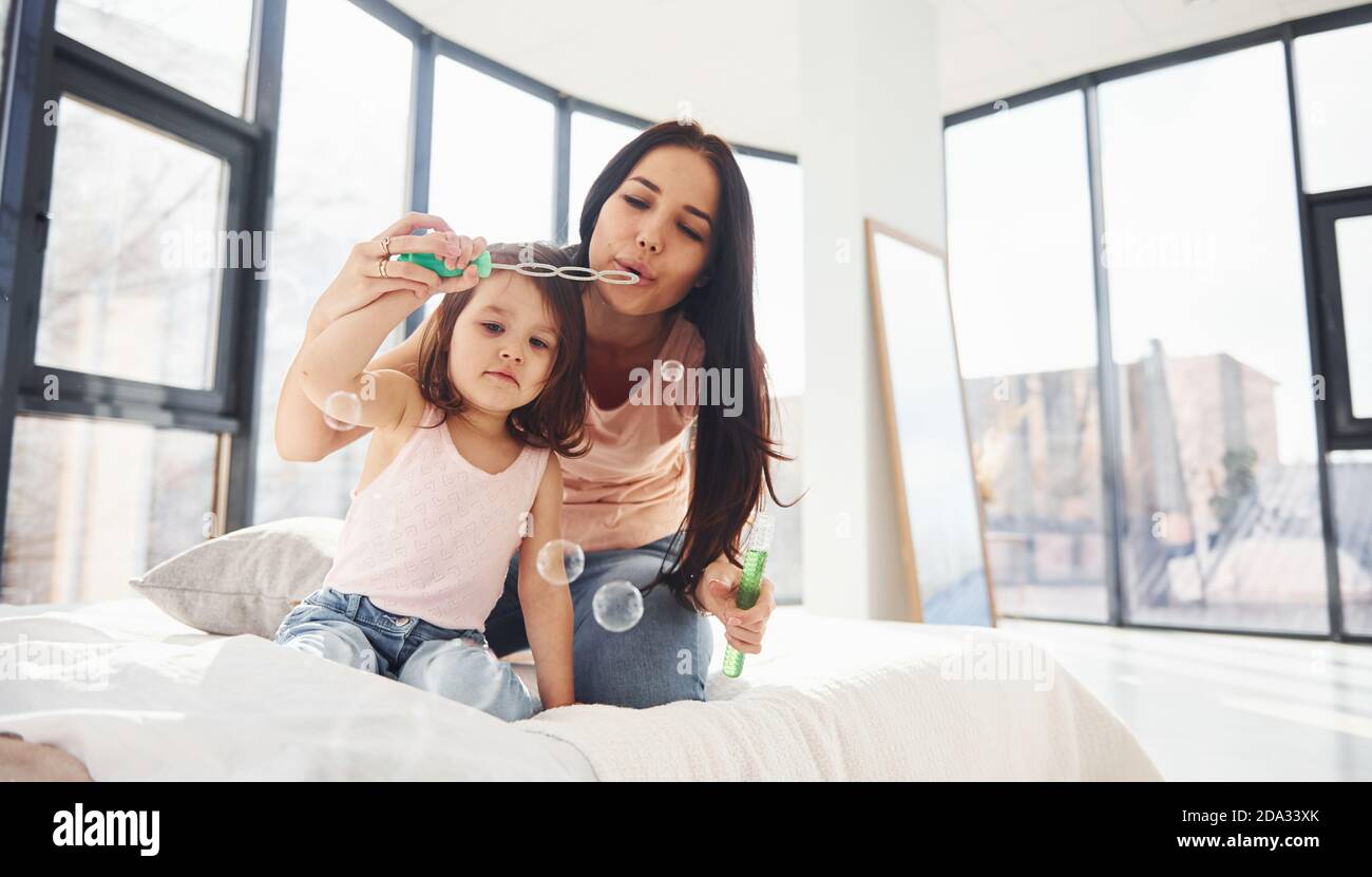 Young mother with her daughter blowing bubbles together in bedroom Stock Photo