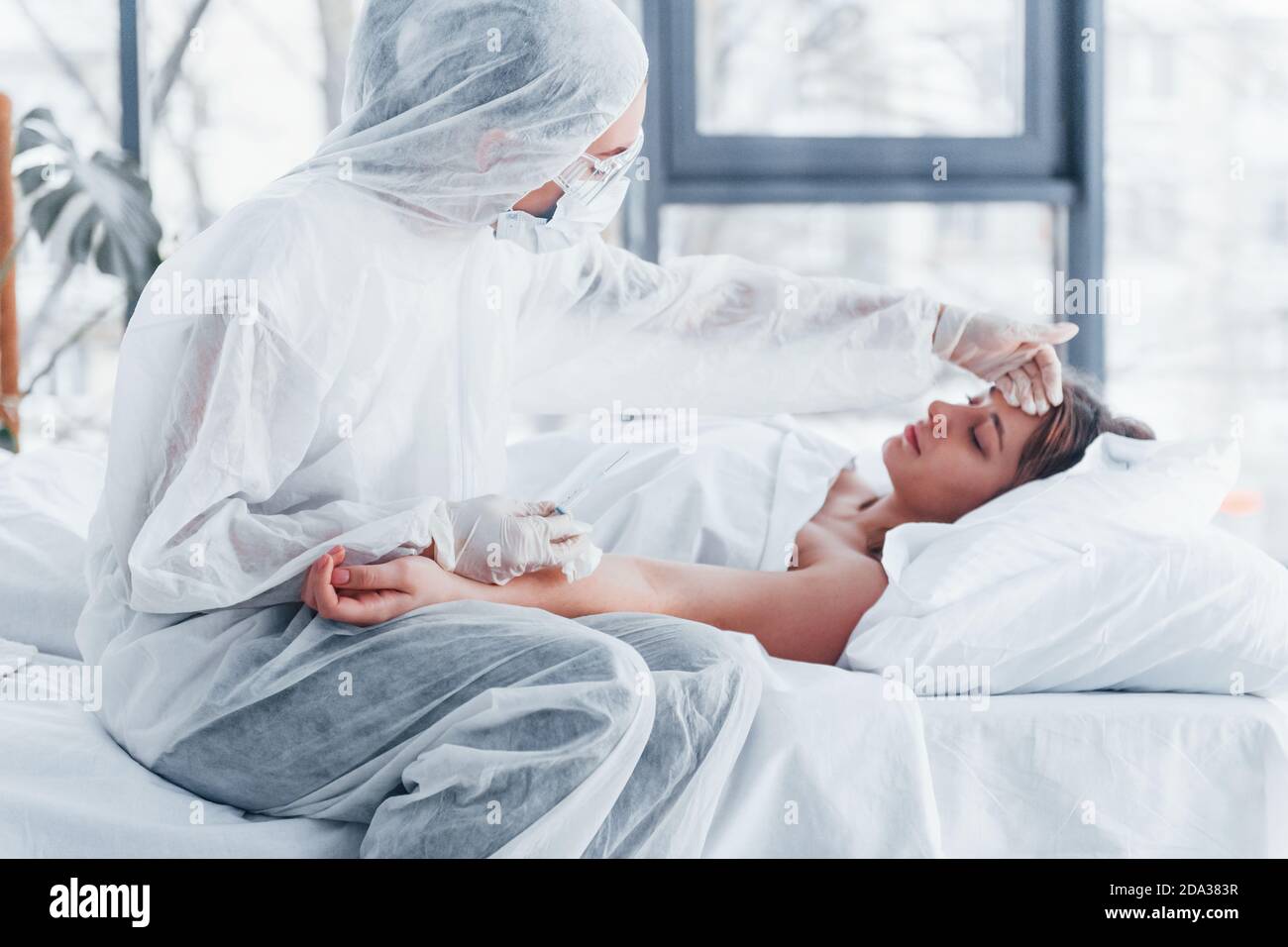Checks temperature. Female doctor in defensive lab coat and protective eyewear with syringe in hand injecting medicine to young girl sick of virus Stock Photo