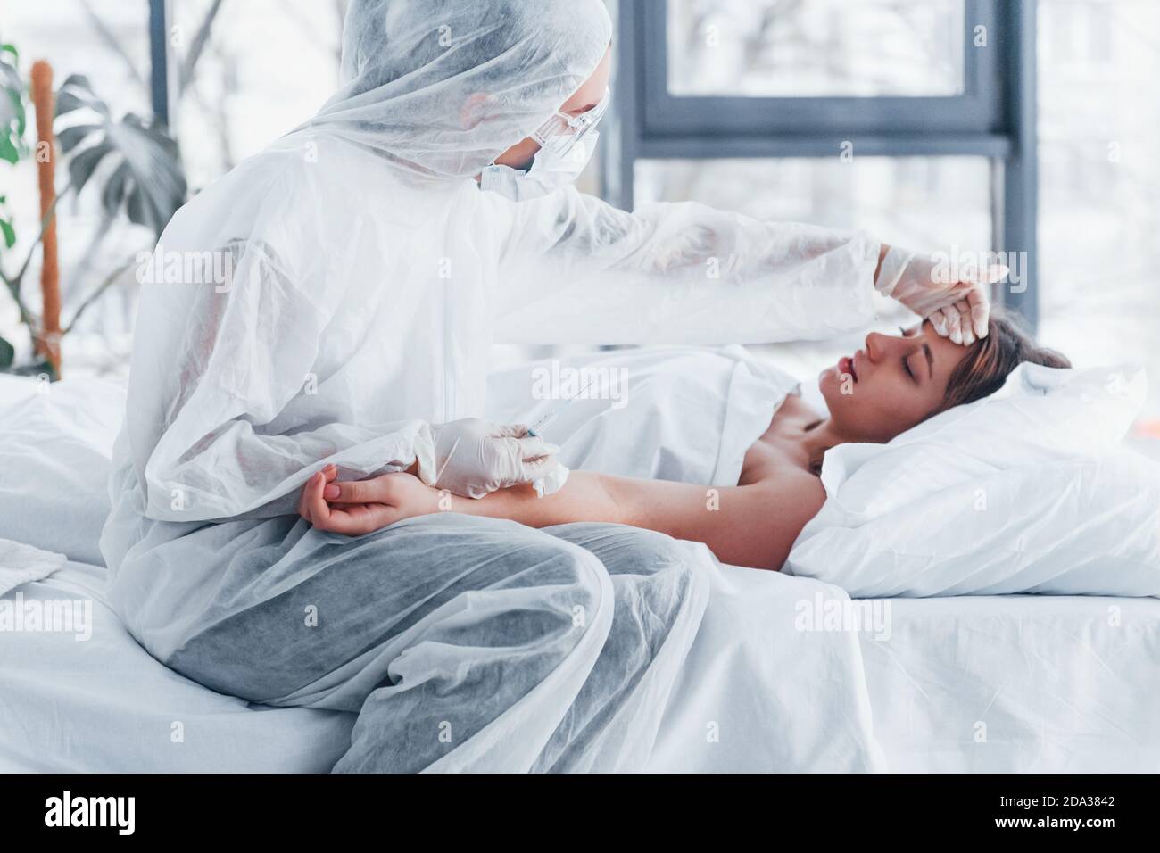 Checks temperature. Female doctor in defensive lab coat and protective eyewear with syringe in hand injecting medicine to young girl sick of virus Stock Photo