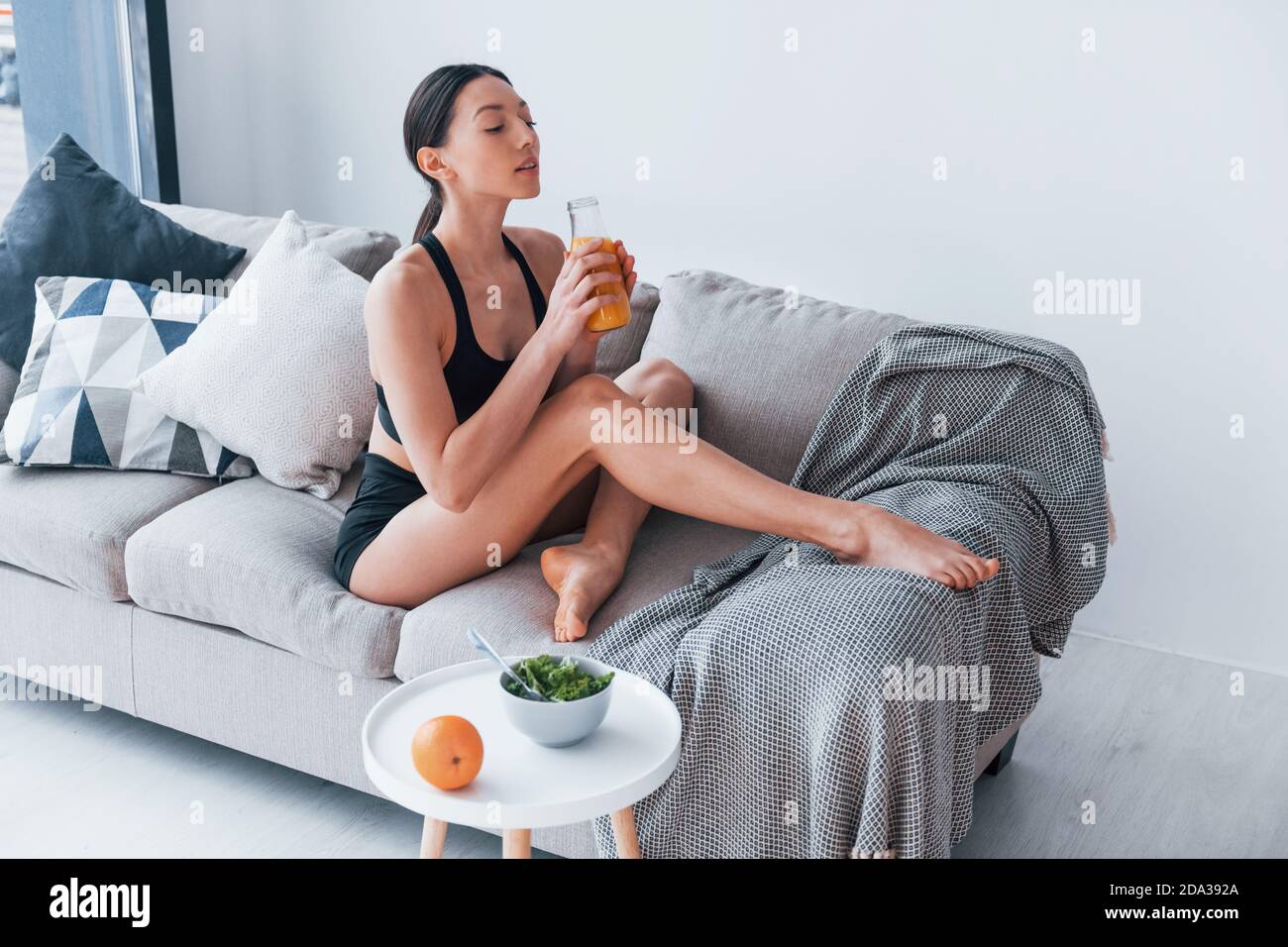Young woman with slim body shape in sportswear sits on sofa and eats healthy diet food indoors at home Stock Photo
