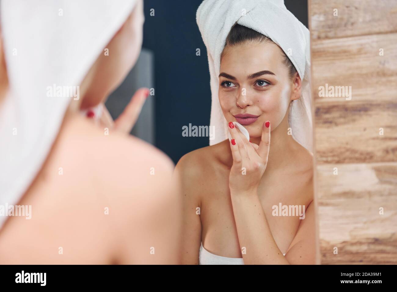 Beautiful young woman standing in bathroom, looking into the mirror and taking care of her face. Stock Photo