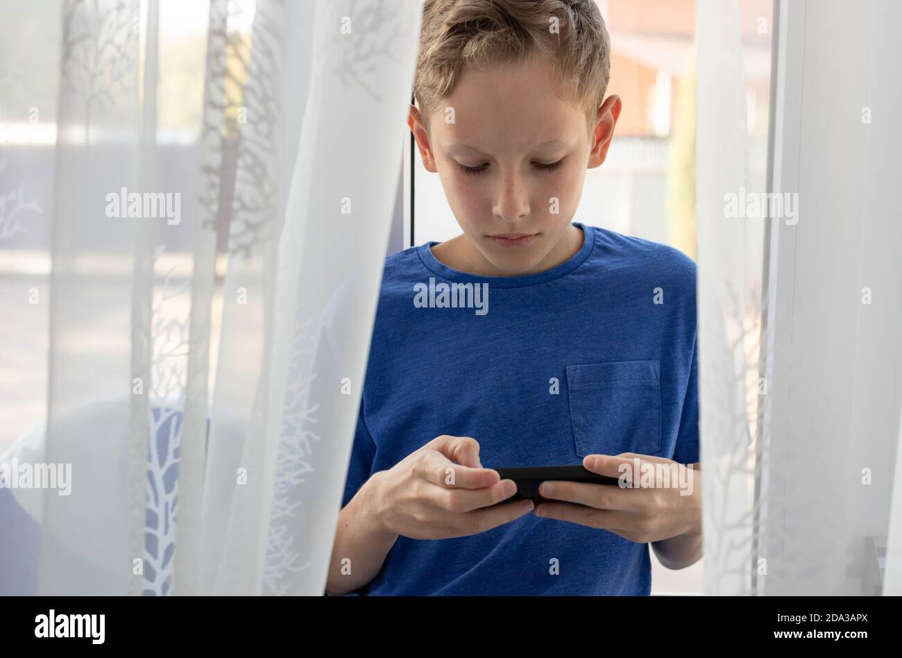 child boy uses mobile phone while sitting on the windowsill at home. Stock Photo
