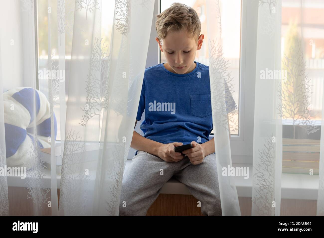 child boy typing message on smartphone while sitting on windowsill at home. Stock Photo