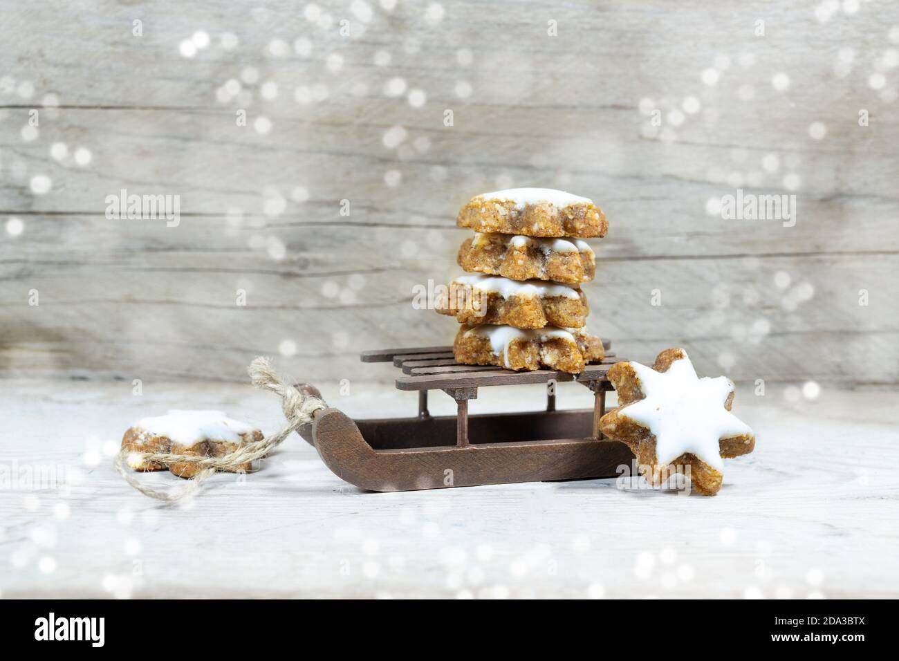 Small sleigh with a stack of cinnamon stars on a rustic grey wooden background with snowy bokeh light spots and copy space, seasonal decoration for Ch Stock Photo