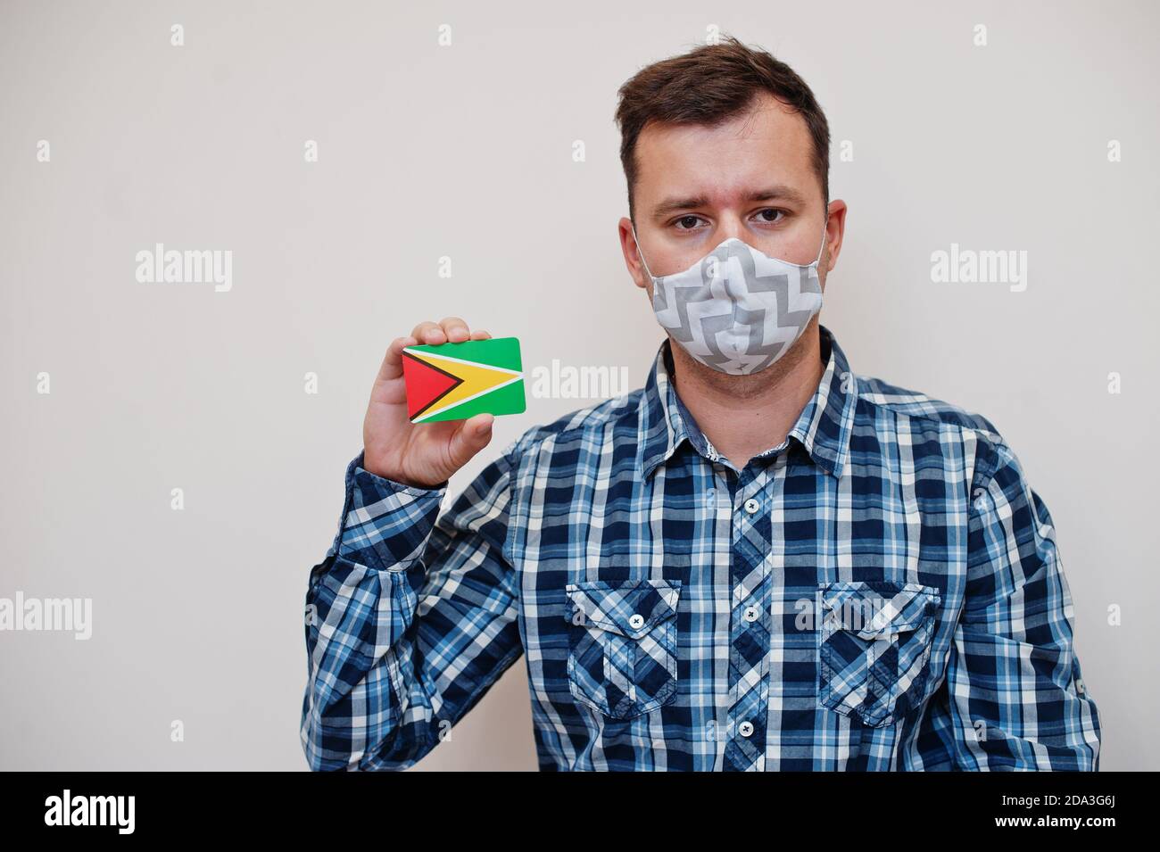 Man in checkered shirt show Guyana flag card in hand, wear protect mask isolated on white background. American countries Coronavirus concept. Stock Photo