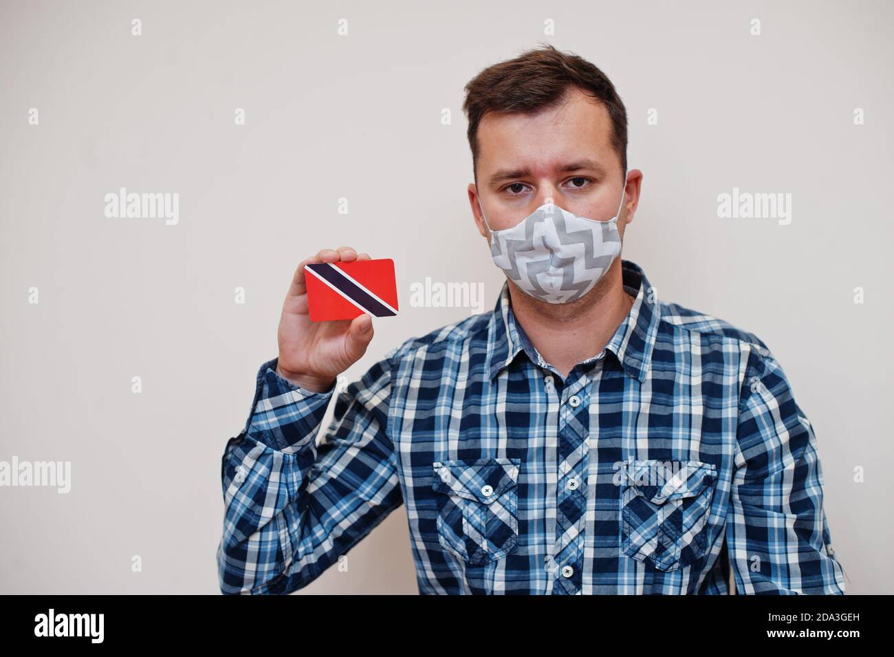 Man in checkered shirt show Trinidad and Tobago flag card in hand, wear protect mask isolated on white background. American countries Coronavirus conc Stock Photo