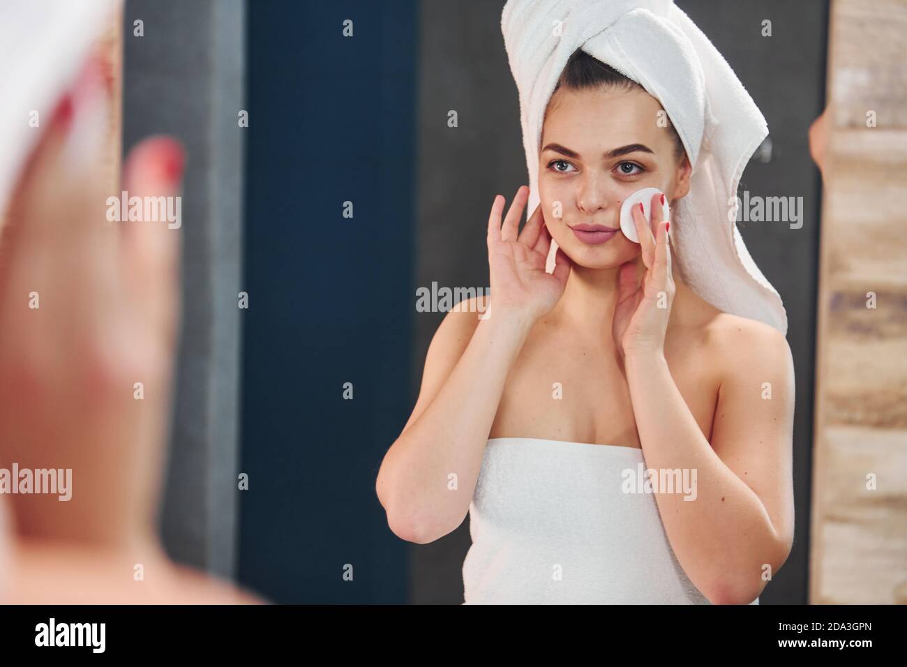 Beautiful young woman standing in bathroom, looking into the mirror and taking care of her face Stock Photo