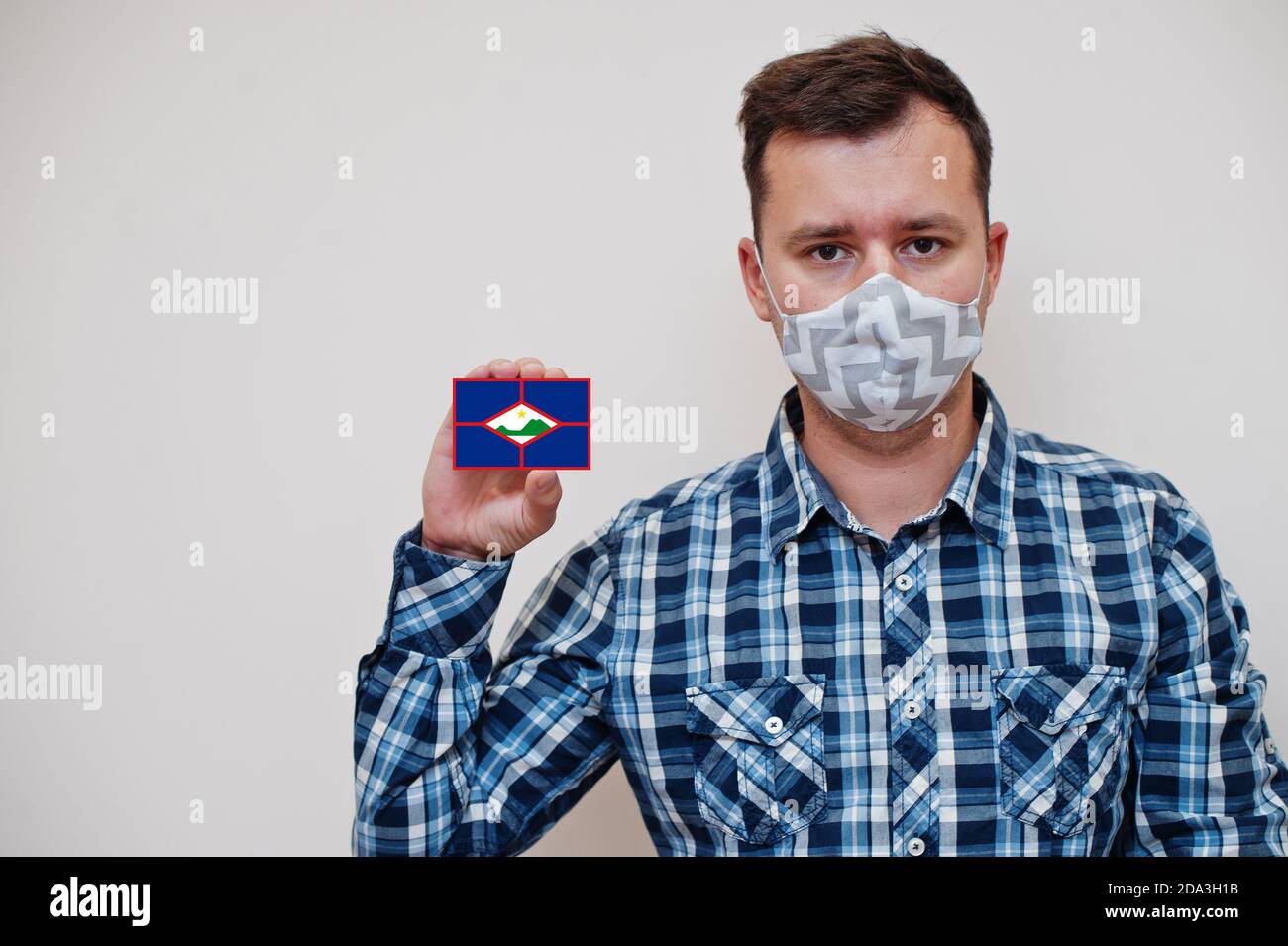 Man in checkered shirt show Sint Eustatius flag card in hand, wear protect mask isolated on white background. American countries Coronavirus concept. Stock Photo