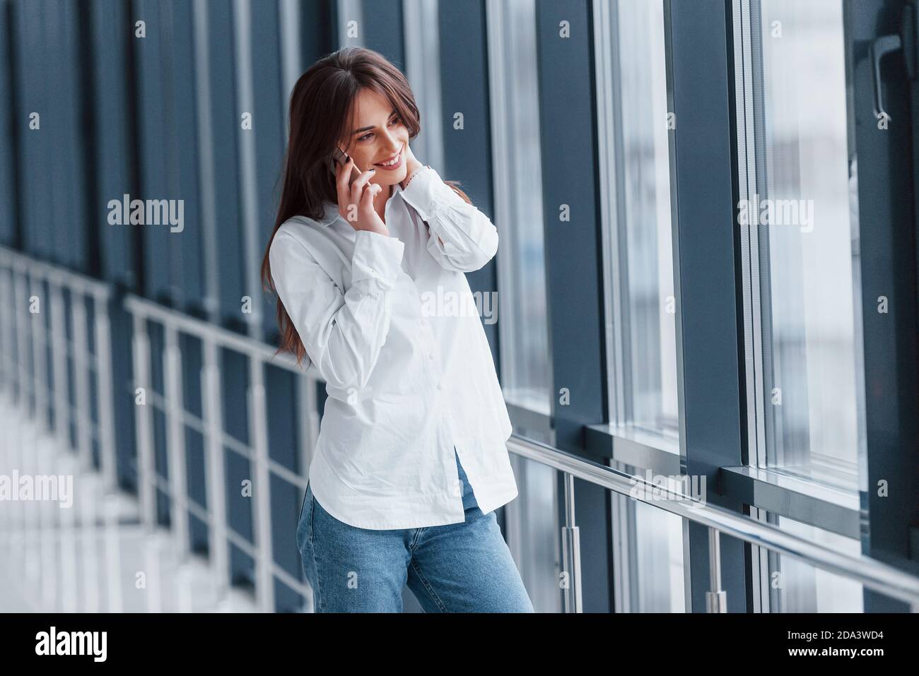 Brunette in white shirt indoors talking by phone in modern airport or hallway at daytime Stock Photo