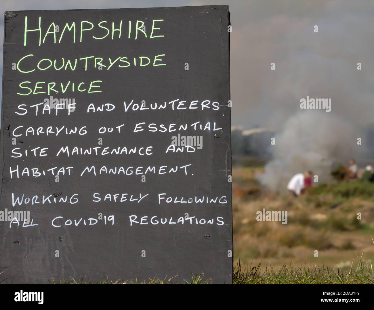 Chalkboard Sign Of Hampshire Countryside Service Warning Of Site Maintenance and Habitat Management Within Covid Regulations With Workers And A Fire I Stock Photo