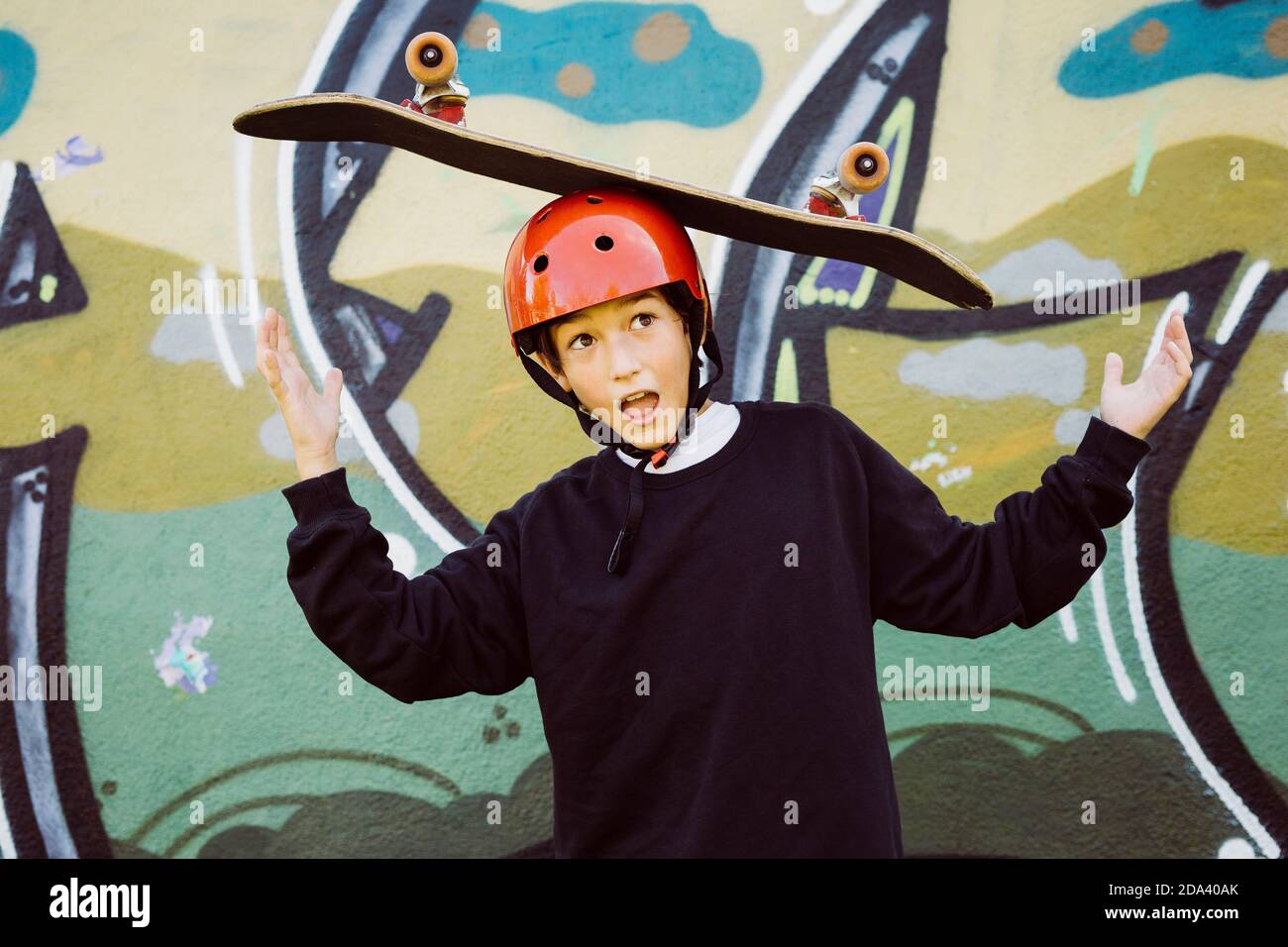 Portrait of a young skater boy wearing  red helmet and playing with an old skateboard, in front of a graffiti mural Stock Photo