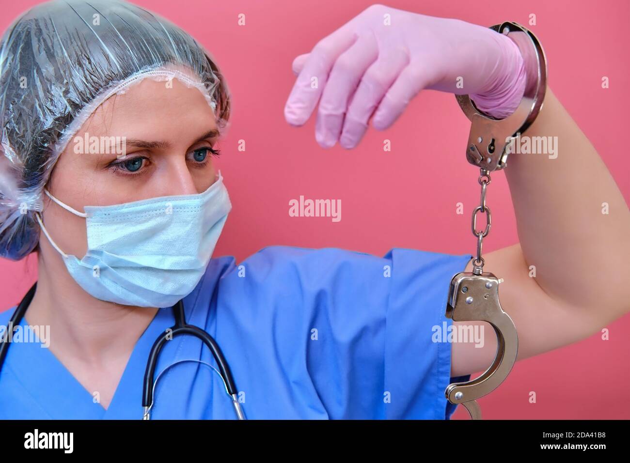 Doctor hands in handcuffs on red background, closeup. Woman hands in pink medical gloves handcuffed, coronavirus quarantine concept. Stock Photo