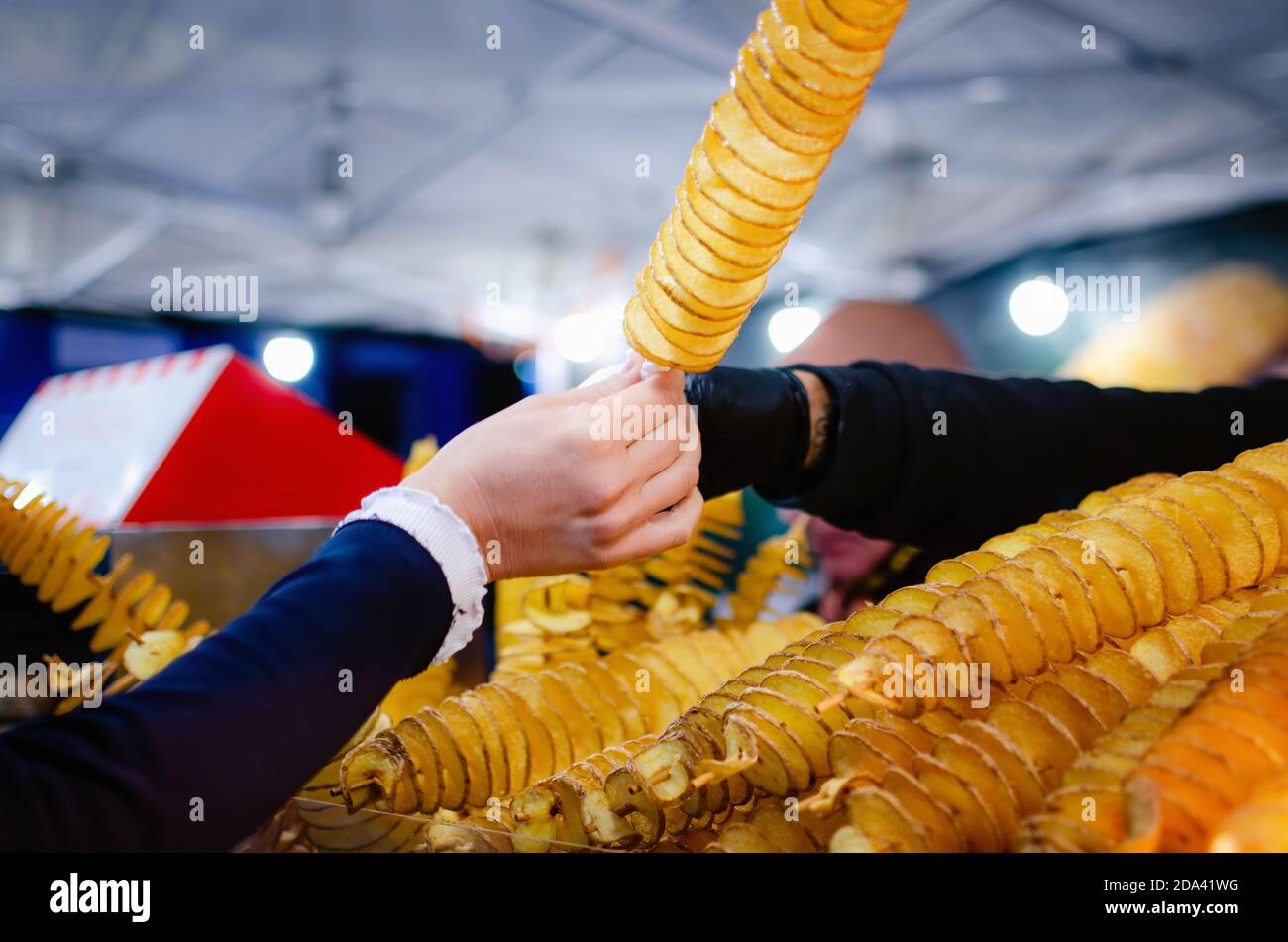 female hand taking a spiral twisted tornado potato from the seller of a street food market stall at evening Stock Photo