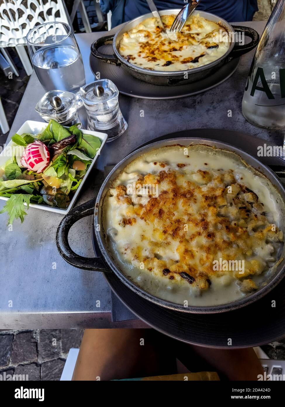 Three cheese crozet pasta with cream in a cast iron skillet and salad on the side and glasses of water. Delicious meal served on a table. Stock Photo