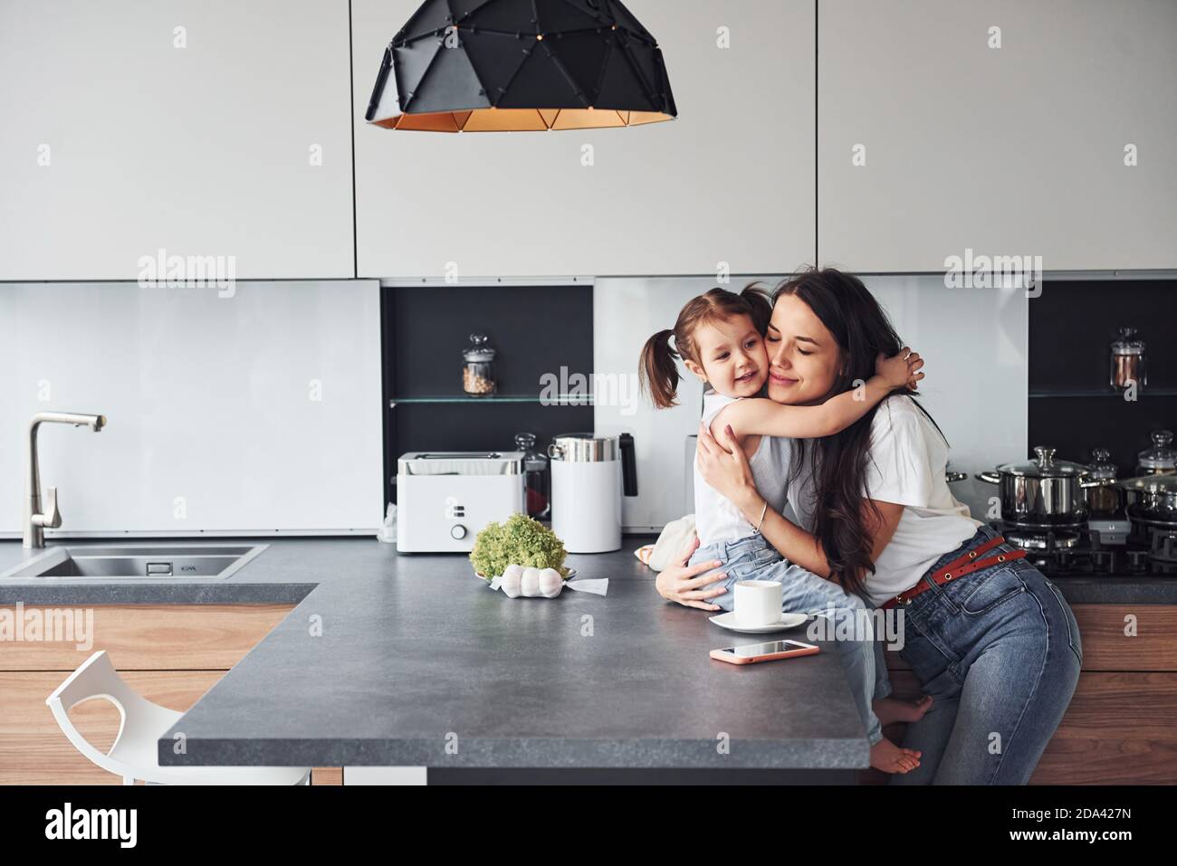 Mother with her little daughter embracing each other indoors in kitchen Stock Photo