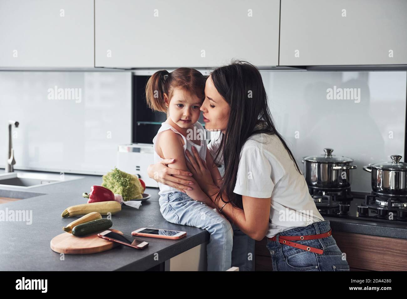 Mother with her little daughter embracing each other indoors in kitchen Stock Photo
