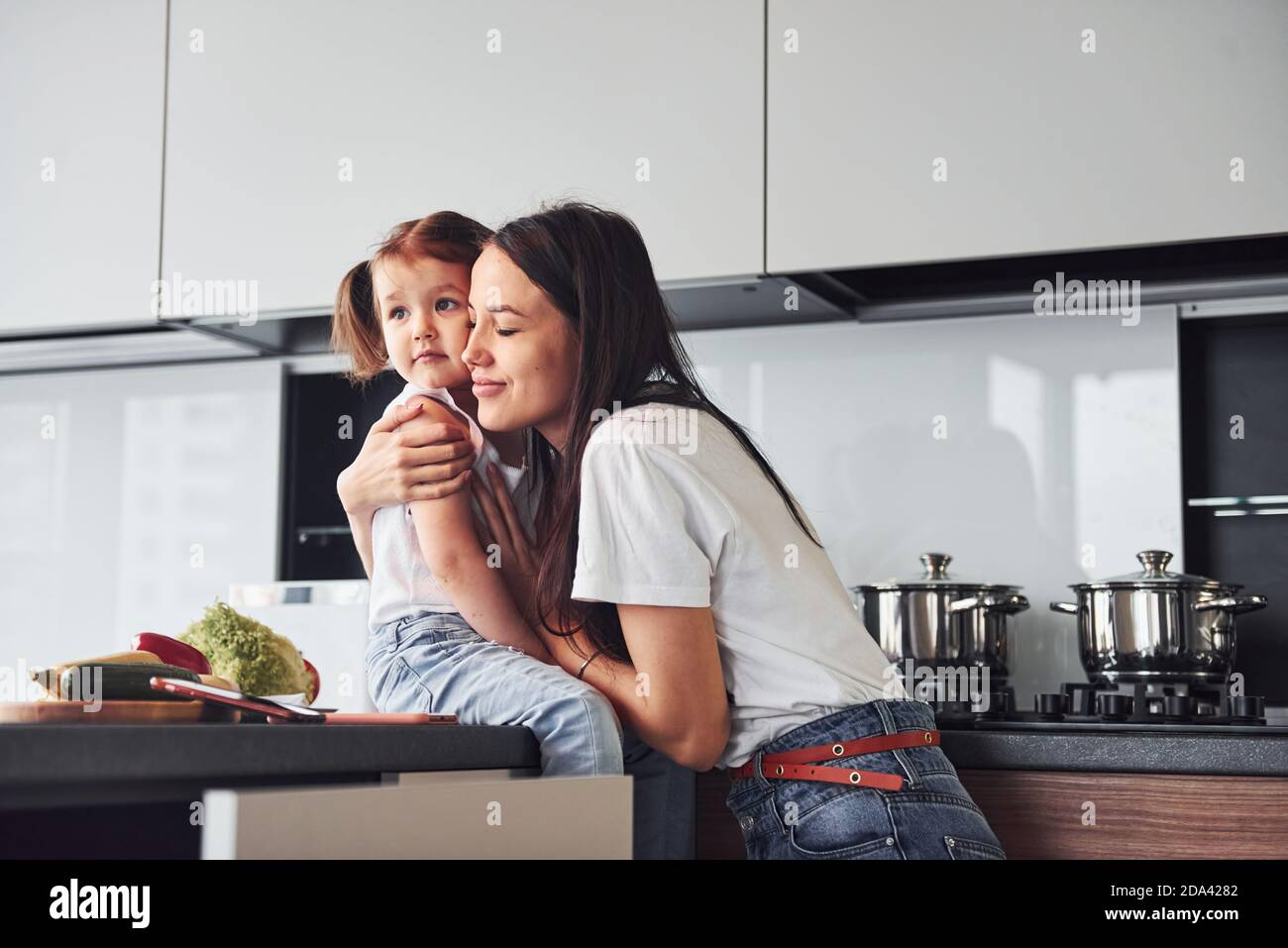 Mother with her little daughter embracing each other indoors in kitchen Stock Photo