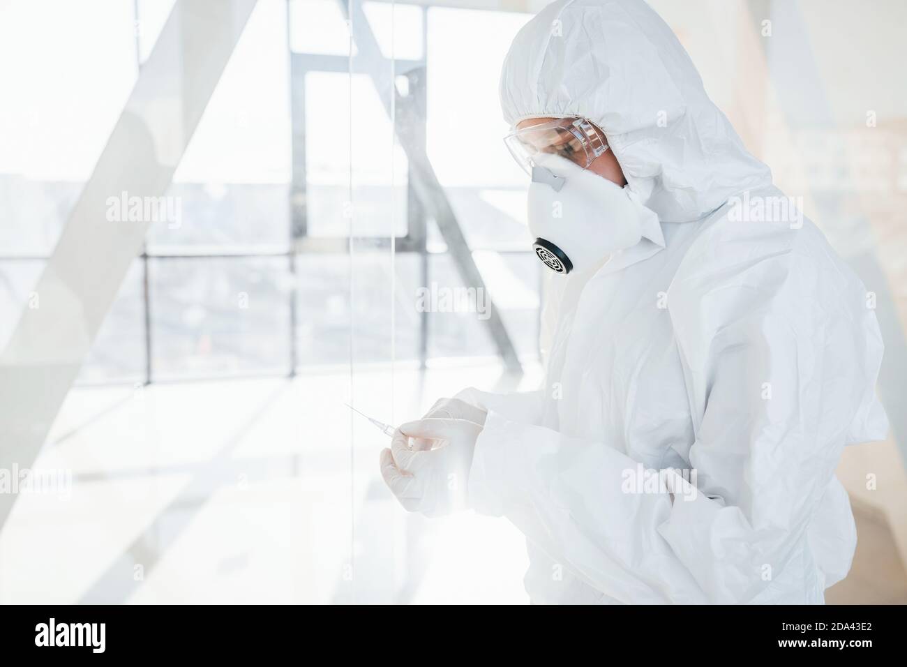 Holds syringe with medicine. Female doctor scientist in lab coat, defensive eyewear and mask standing indoors Stock Photo