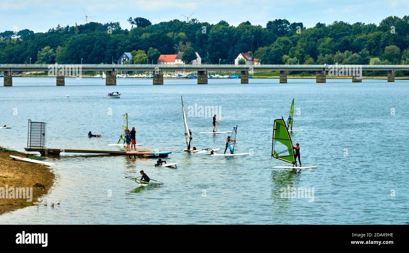 Moehnesse, Germany, July 22., 2020: Group of teenagers practice surfing on windsurfing boards in the lake with a long bridge in the background Stock Photo