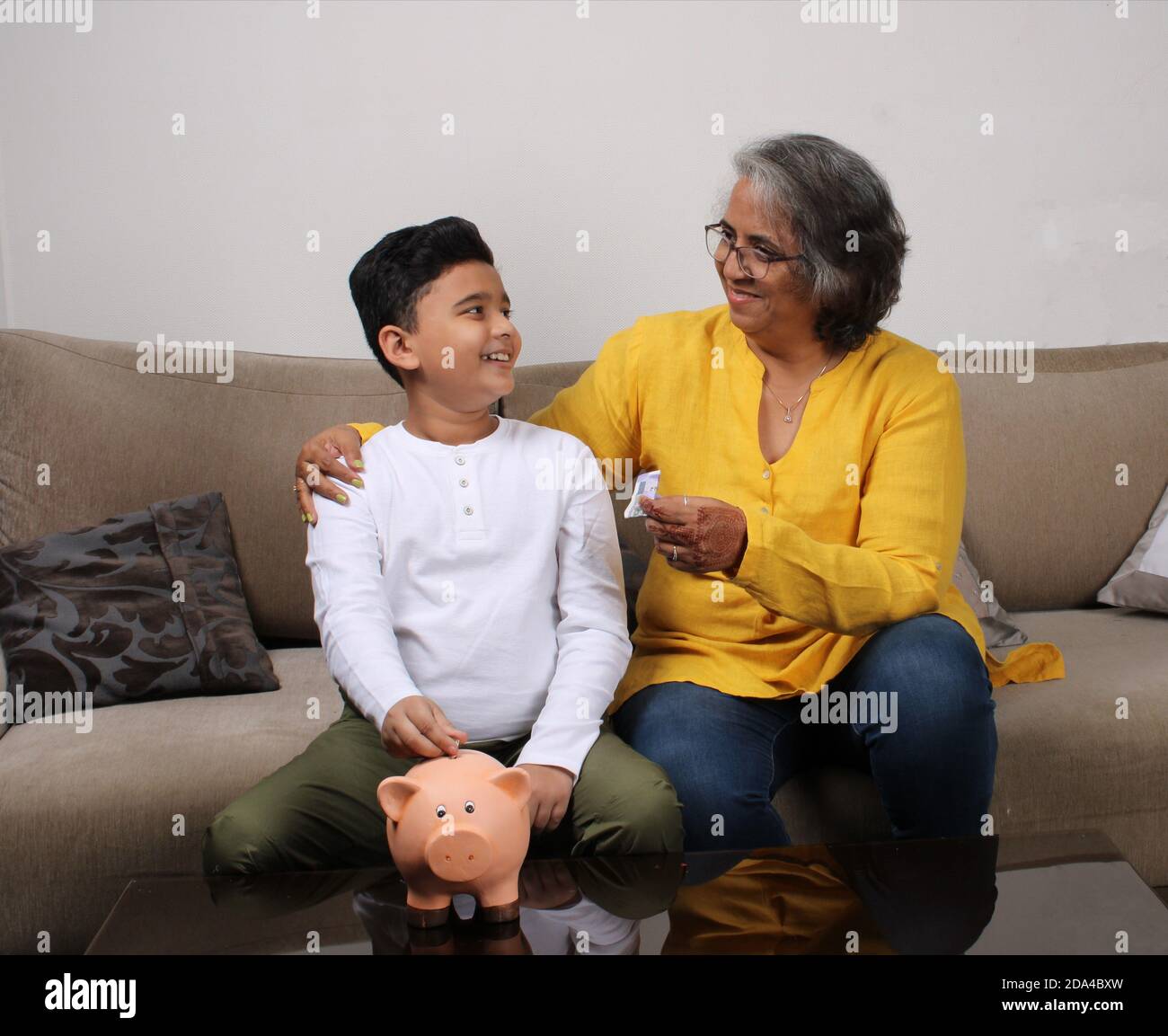 Indian/asian Grandma teaching her grand son importance of savings, putting coins into pink piggy bank. Stock Photo