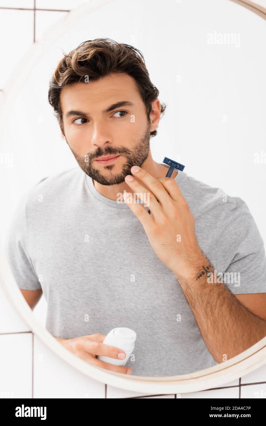 Bearded man touching chin while holding razor and shaving foam near
