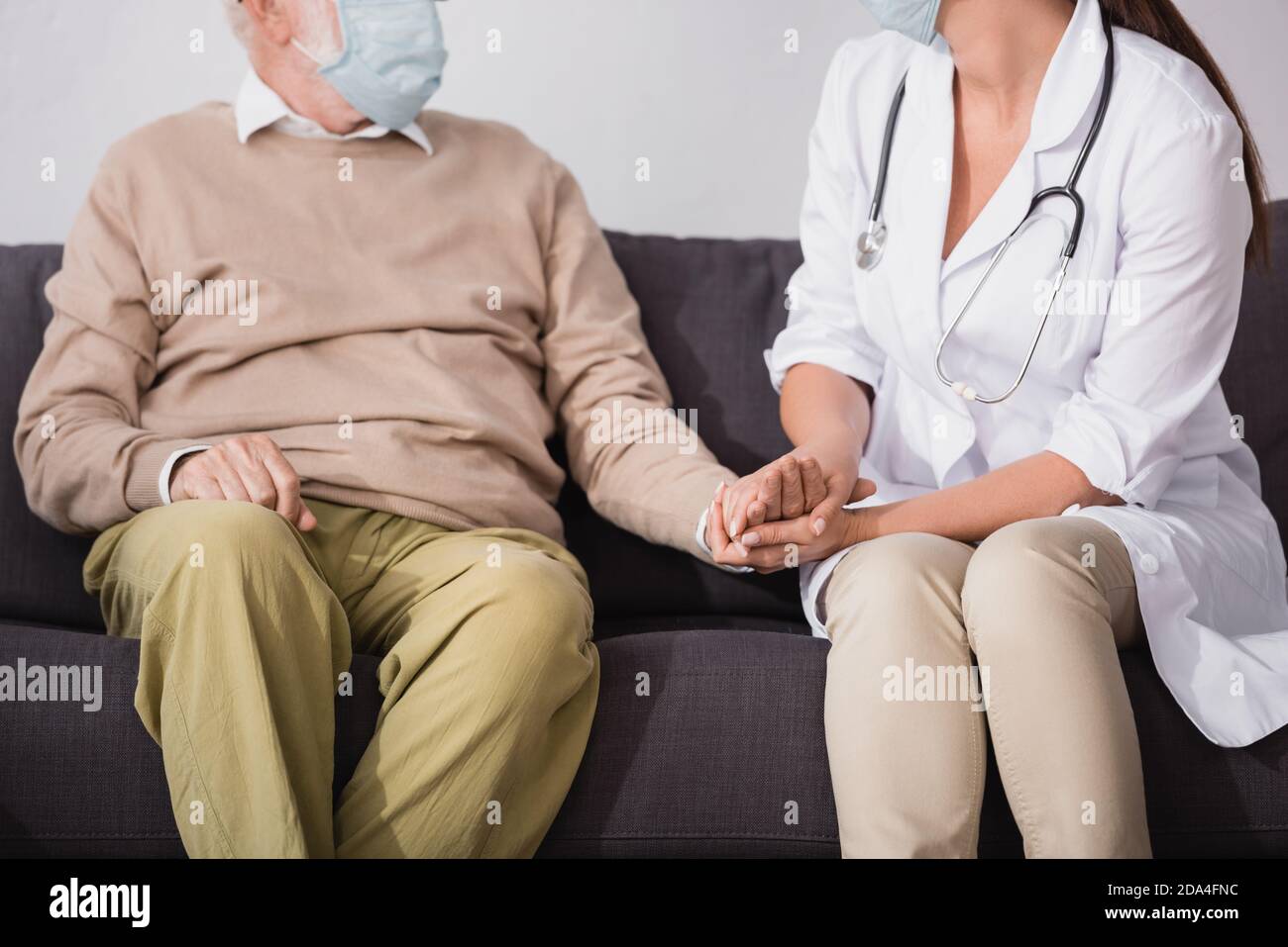 partial view of social worker in medical mask holding hands of aged man at home Stock Photo