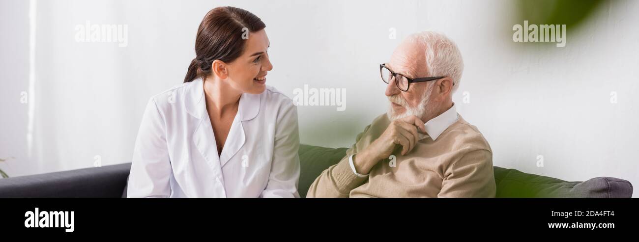 geriatric nurse and aged man talking while sitting on sofa at home, banner Stock Photo