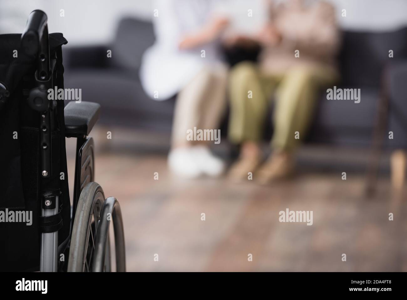 medical assistant and aged man sitting on sofa at home near wheelchair on blurred foreground Stock Photo