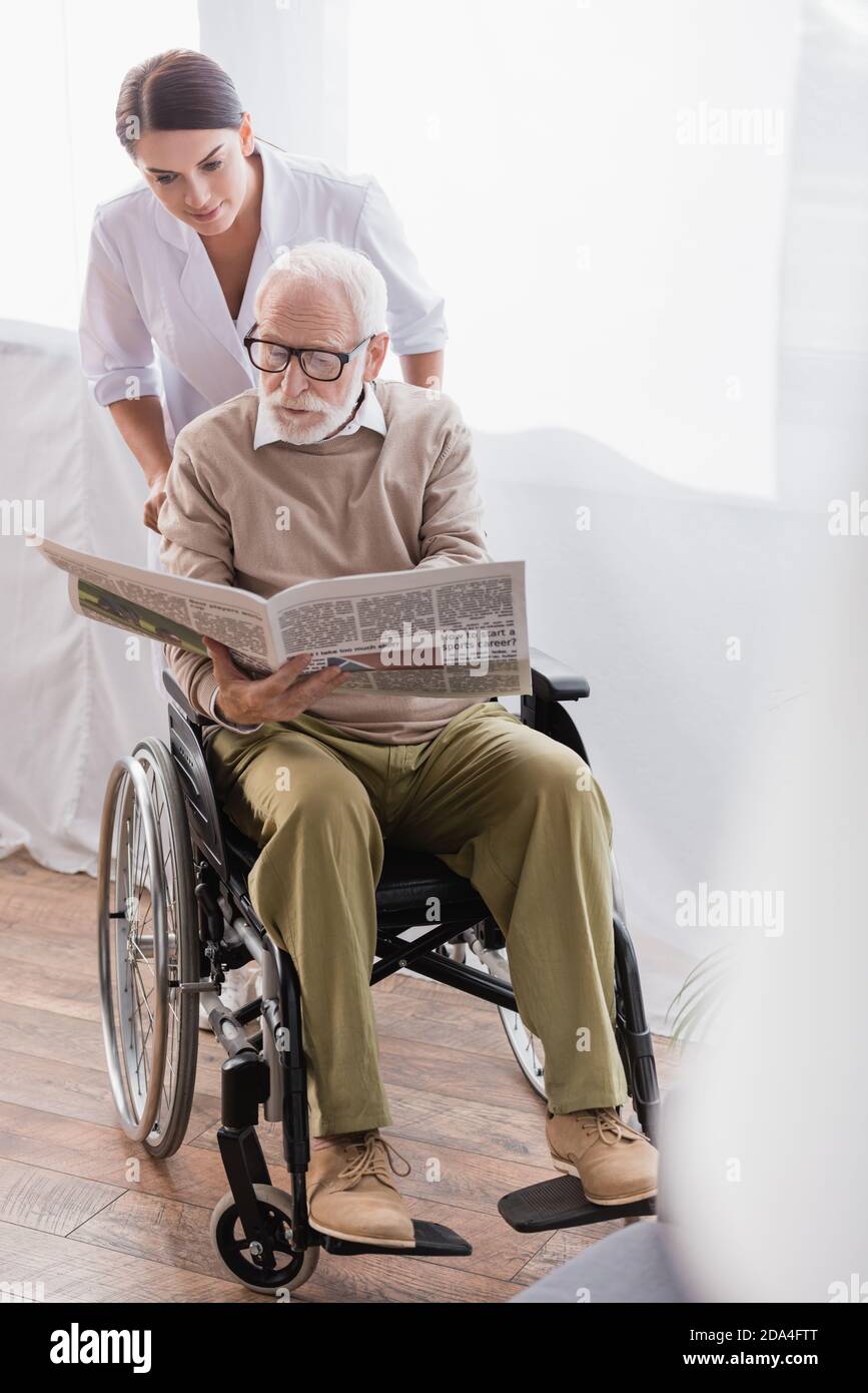 aged disabled man reading newspaper in wheelchair near social worker Stock Photo