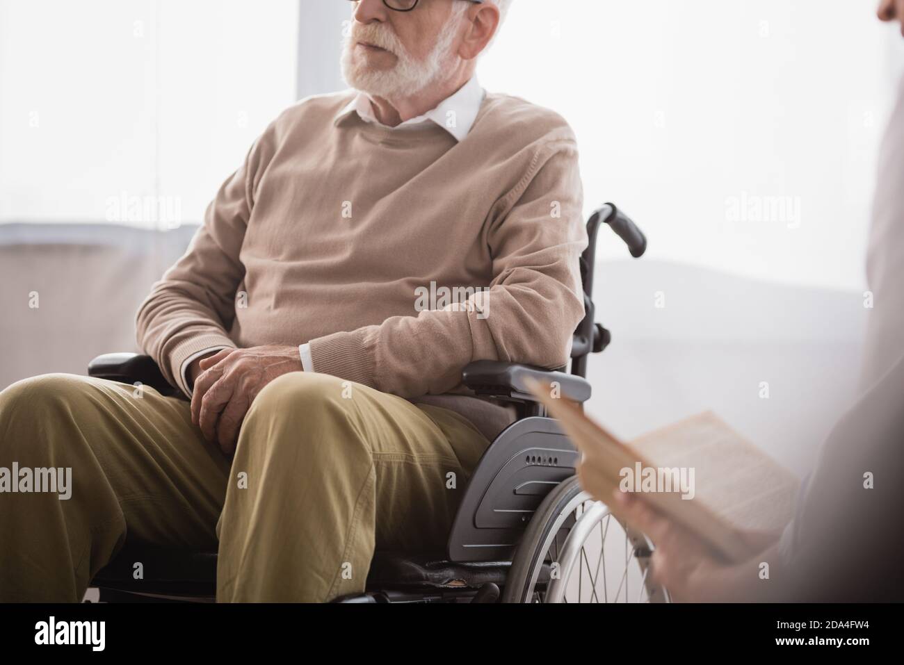 cropped view of social worker reading book to disabled man in wheelchair on blurred foreground Stock Photo