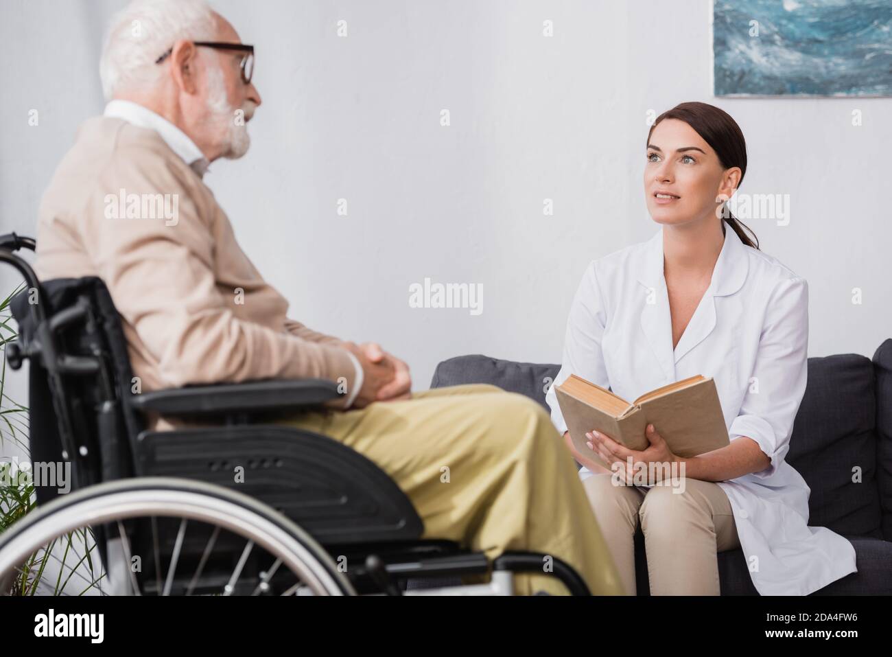 social worker in white coat reading book to aged disabled man in wheelchair on blurred foreground Stock Photo