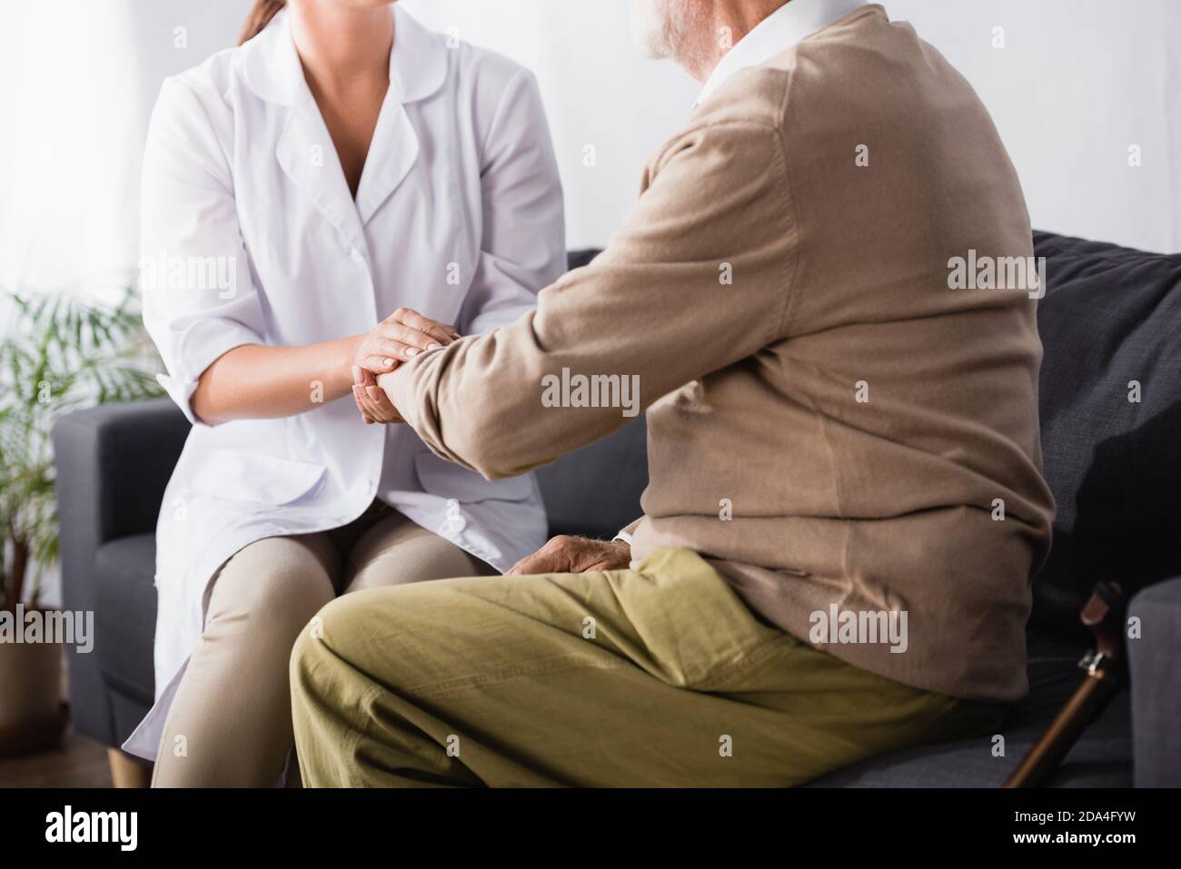 partial view of aged man and geriatric nurse holding hands while sitting on sofa Stock Photo