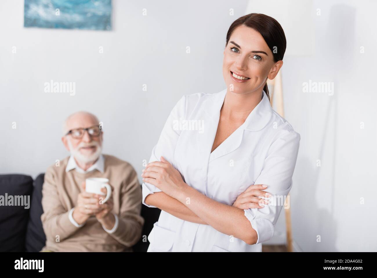 cheerful social worker smiling at camera while standing with crossed arms near aged man holding cup of tea Stock Photo