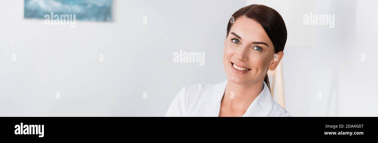 cheerful brunette nurse in white coat smiling at camera, banner Stock Photo