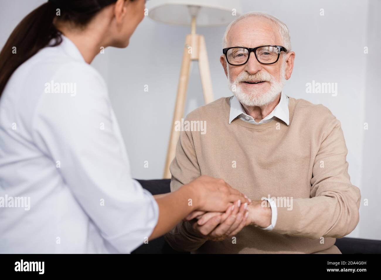 elderly man looking at camera while holding hands with social worker at home on blurred foreground Stock Photo