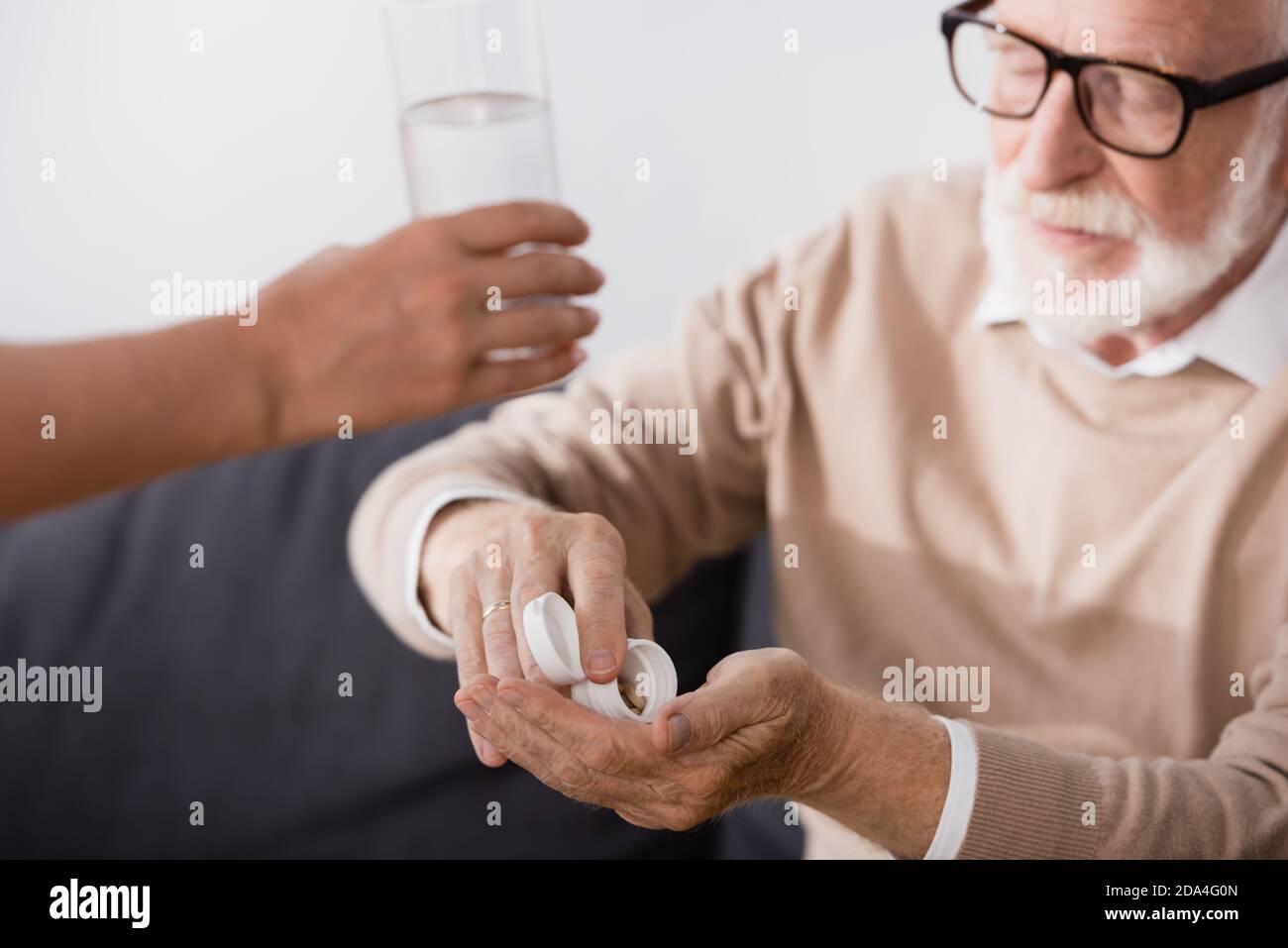elderly man in eyeglasses taking pills near nurse giving glass of water on blurred foreground Stock Photo