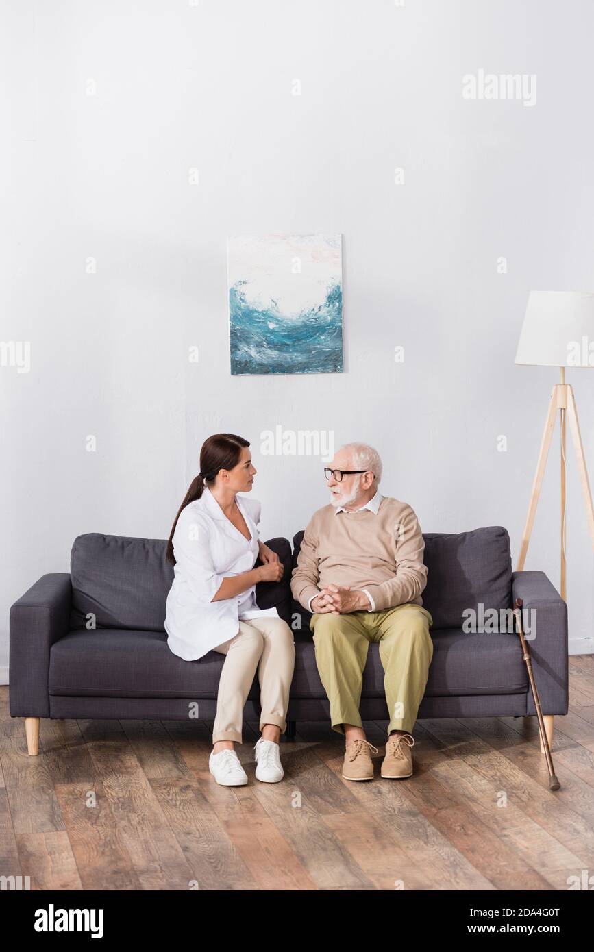 aged man in eyeglasses talking to geriatric nurse while sitting on sofa at home Stock Photo