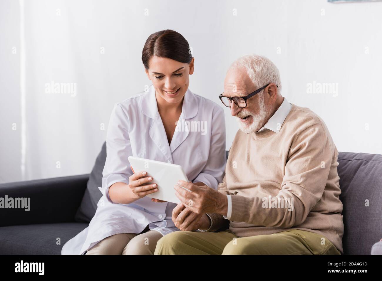 brunette social worker showing digital tablet to smiling aged man at home Stock Photo