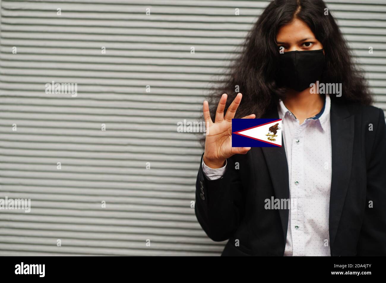 Asian woman at formal wear and black protect face mask hold American Samoa flag at hand against gray background. Coronavirus at country concept. Stock Photo
