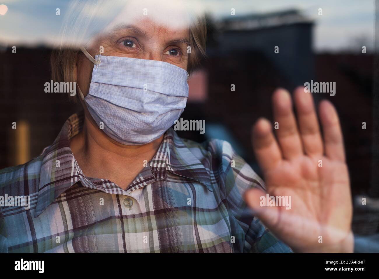 Elderly caucasian woman wearing hand made protective face mask, in nursing care home, looking outside window with sadness in her eyes, self isolation Stock Photo