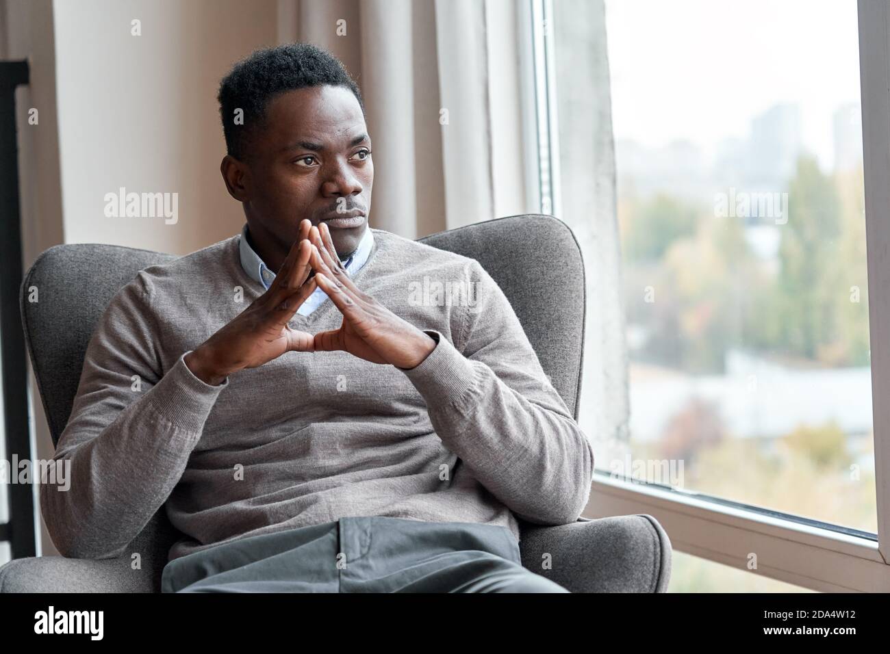 Confident stylish black man sitting in chair at home looking through window. Stock Photo