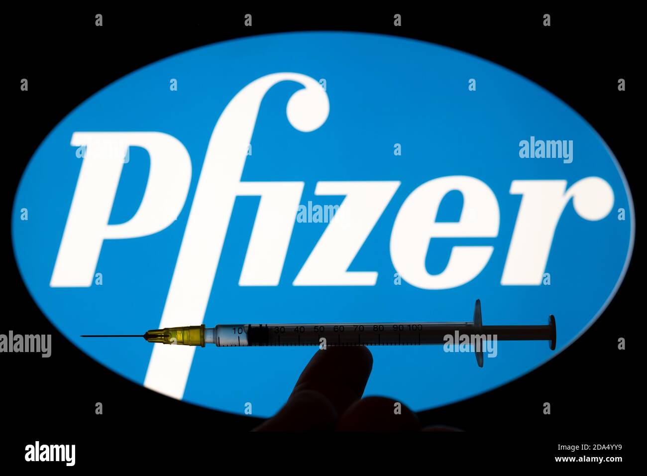 Stafford / United Kingdom - November 9 2020: Pfizer Covid-19 vaccine concept. Syringe balanced on a fingertip and blurred Pfizer company logo on the b Stock Photo