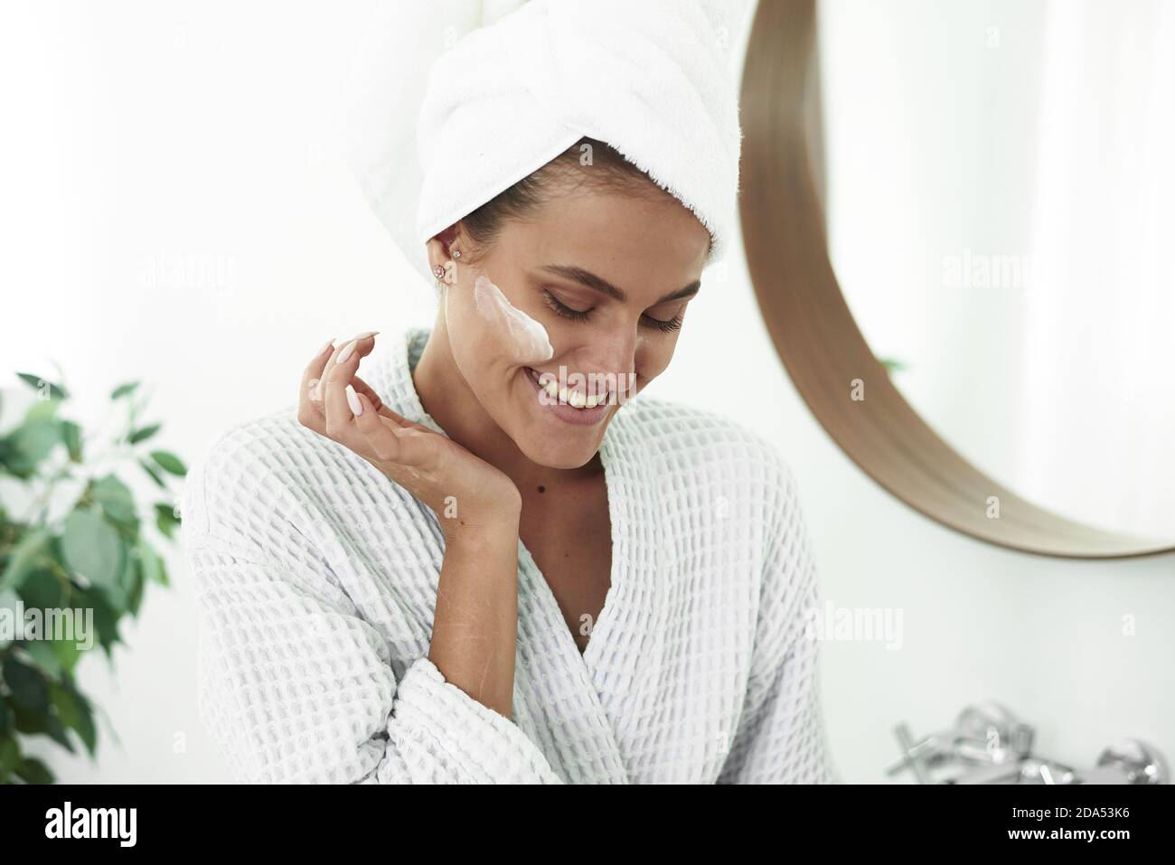 A smiling woman in a bathrobe and a towel on her head applies moisturizer from a jar to her face in the bathroom. The concept of skin care, moisturizing and cleansing the face and neck. Stock Photo