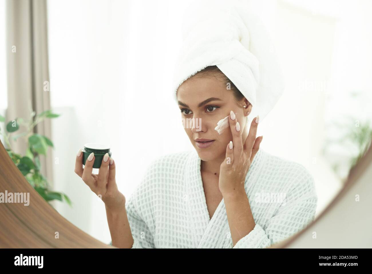 A beautiful young woman with vitiligo on her hands in a bathrobe and with a towel on her head applies moisturizer to her face. The concept of skin care, moisturizing and cleansing the face and neck. Stock Photo