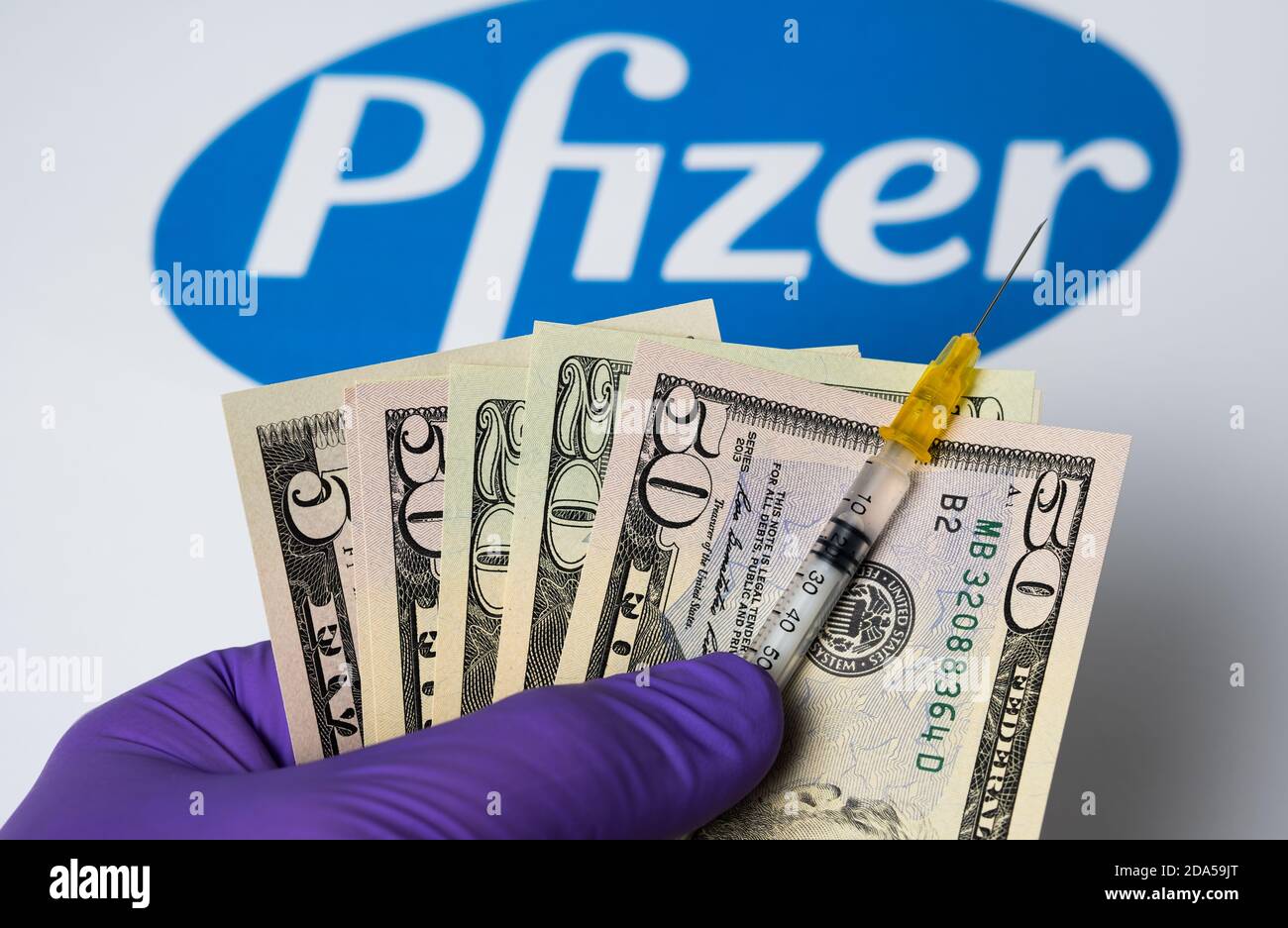 Stafford / United Kingdom - November 9 2020: Pfizer Covid-19 vaccine concept. Syringe with dollars hold in hand and blurred Pfizer company logo on the Stock Photo