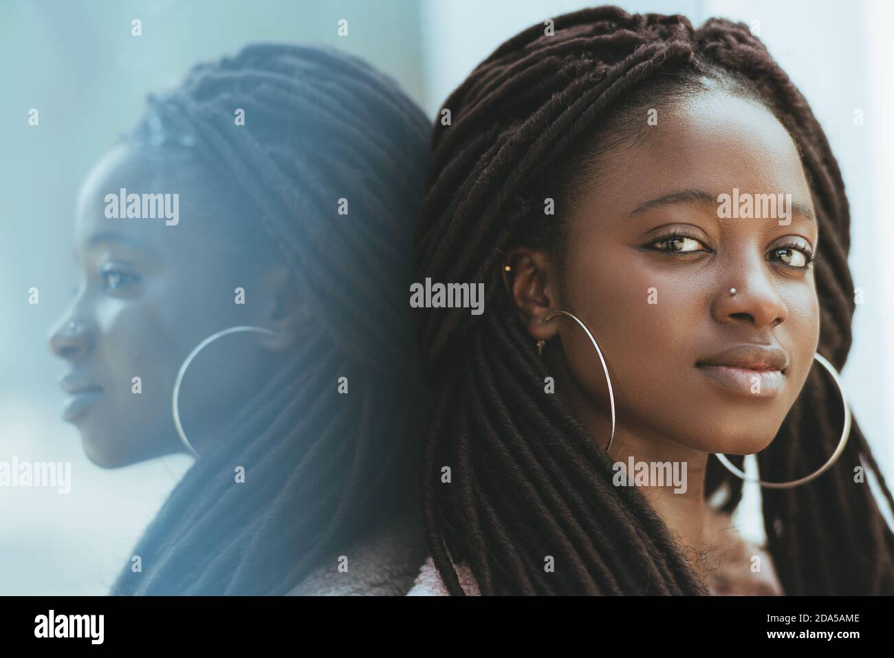 A close-up portrait of a cute young black woman with dreadlocks and huge earrings, piercing in her nose, she is leaning against a glass wall outdoors Stock Photo