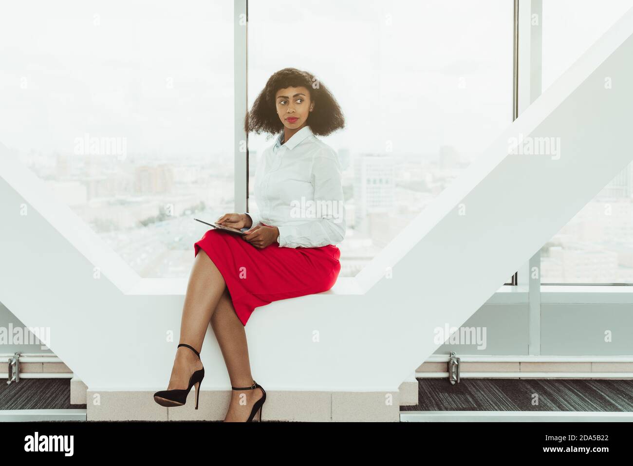 Young ravishing slender African-American businesswoman is sitting on a contemporary window bench of the modern office with a digital tablet in hands a Stock Photo