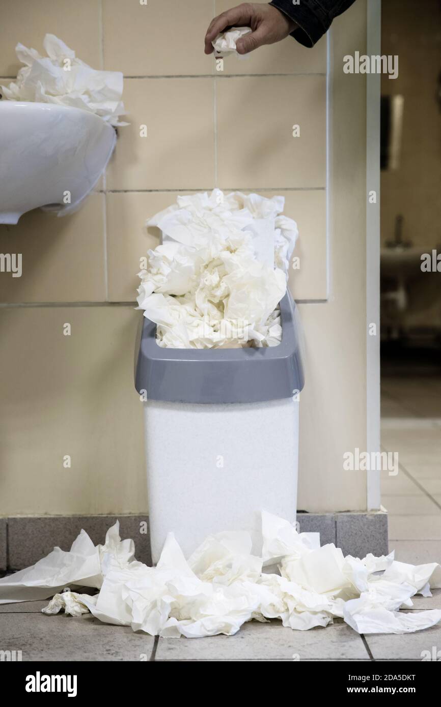 White used paper tissue in overcrowded bin/Male hand drops paper tissue in full trash bin/utilized toilet paper dropped around the trash basket, verti Stock Photo
